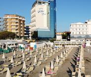 Spiaggia soleggiata con ombrelloni chiusi, hotel moderno sullo sfondo.