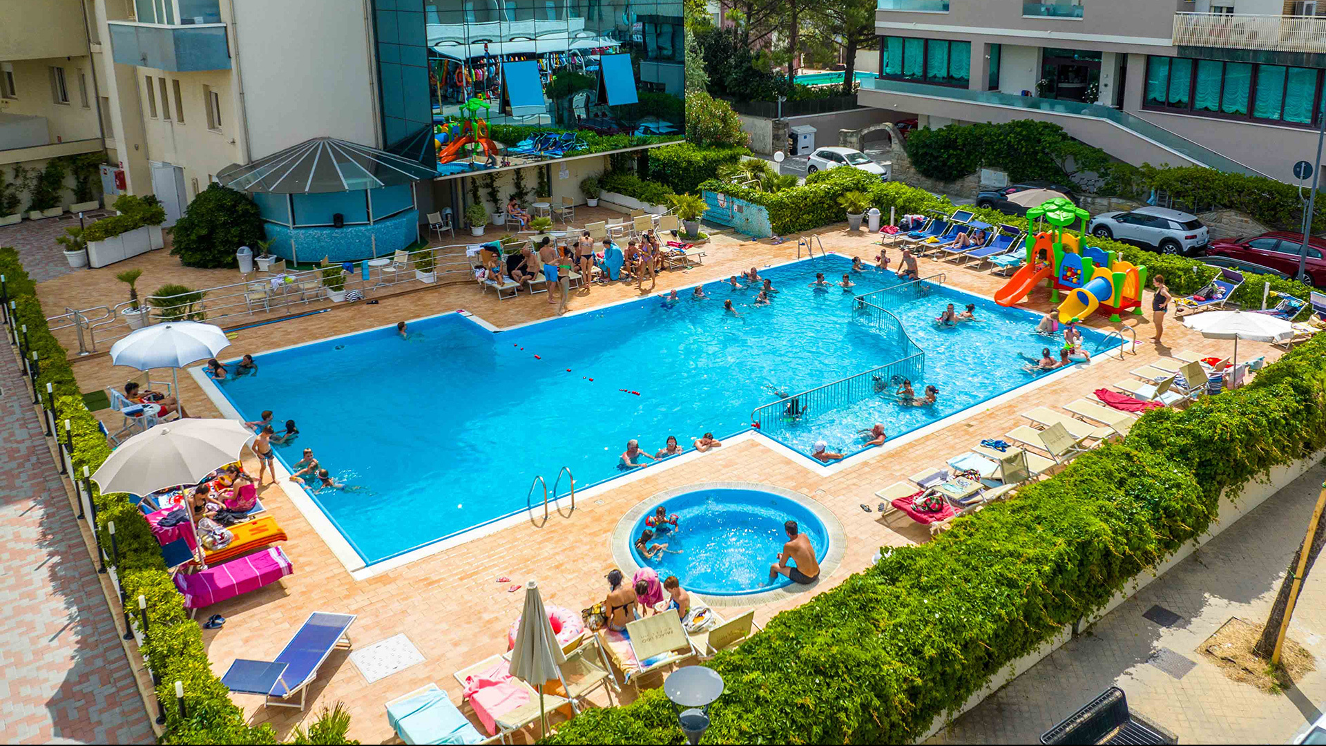 Bambino e uomo in piscina con braccioli, ombrelloni colorati sullo sfondo.