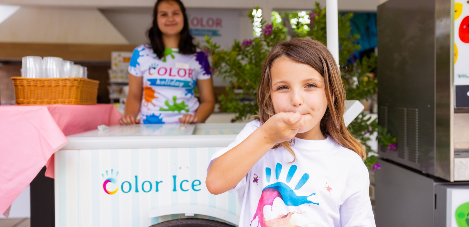 Bambina felice mangia gelato, sorridente venditrice sullo sfondo, giornata di colori vivaci.