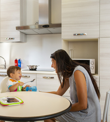Famiglia in cucina, madre prepara, bambino sorride, altra donna dà da mangiare al piccolo.