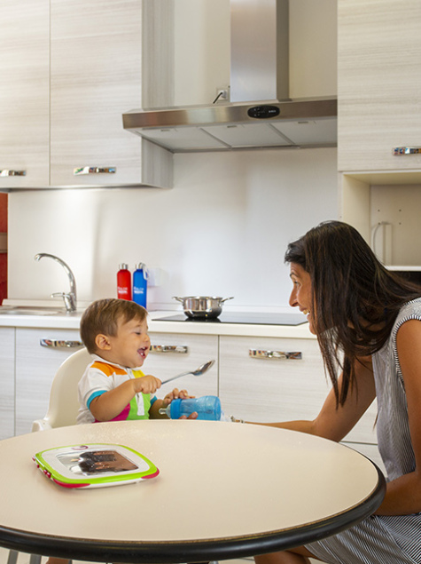 Famiglia in cucina, madre prepara, bambino sorride, altra donna dà da mangiare al piccolo.