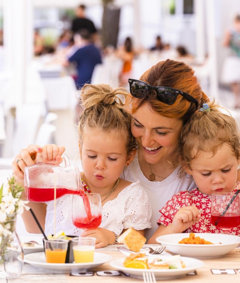 Madre e figlie sorridenti condividono un pasto, versando succo con gioia e affetto.