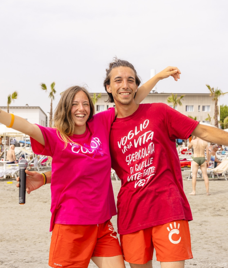 Due amici sorridenti in spiaggia, indossano magliette colorate e trasmettono gioia.